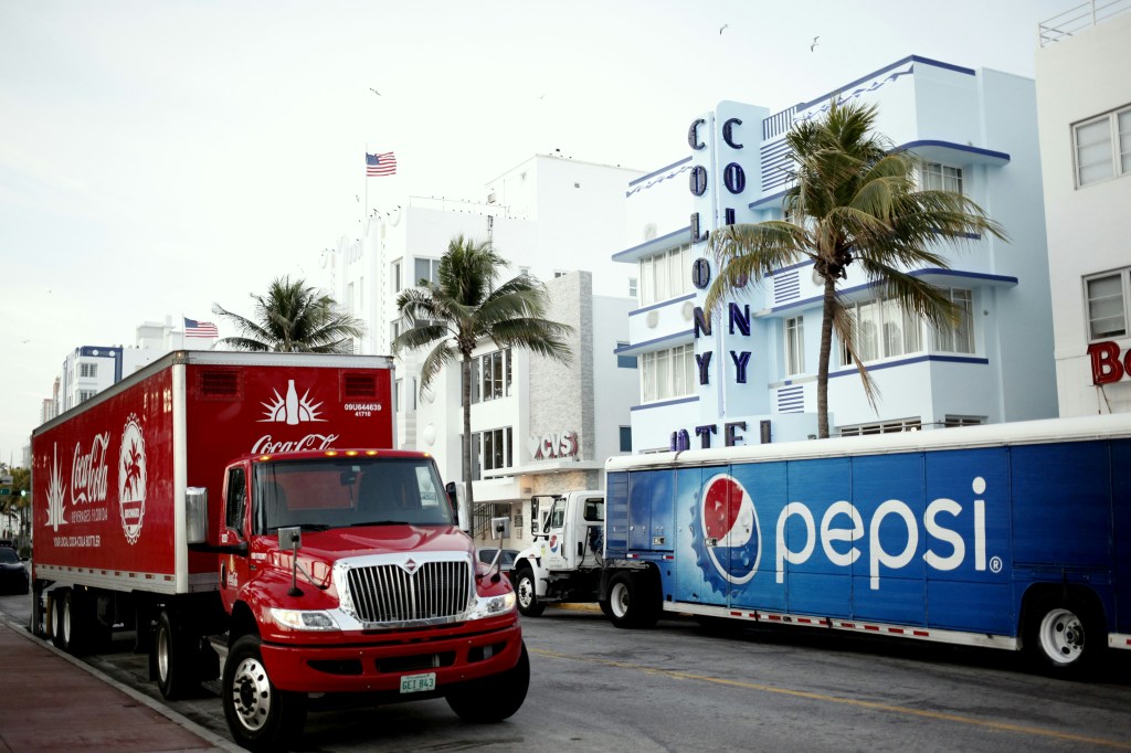 A street view featuring a red Coca-Cola delivery truck parked in front of the Colony Hotel, with a blue Pepsi truck beside it and palm trees in the background.